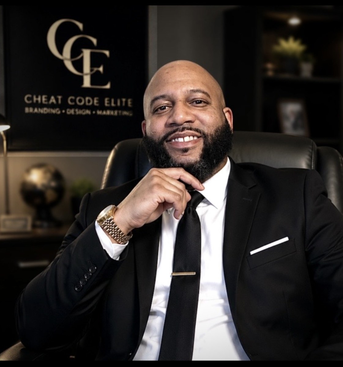 Torey Bush, Founder and Creative Designer of Cheat Code Elite, seated in front of the CCE logo